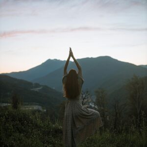 A woman in a dress performs tree pose against a mountainous sunrise backdrop, adding serenity and strength.