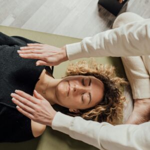 A woman receiving Reiki therapy with hands hovering over her, eyes closed, in a serene indoor setting.